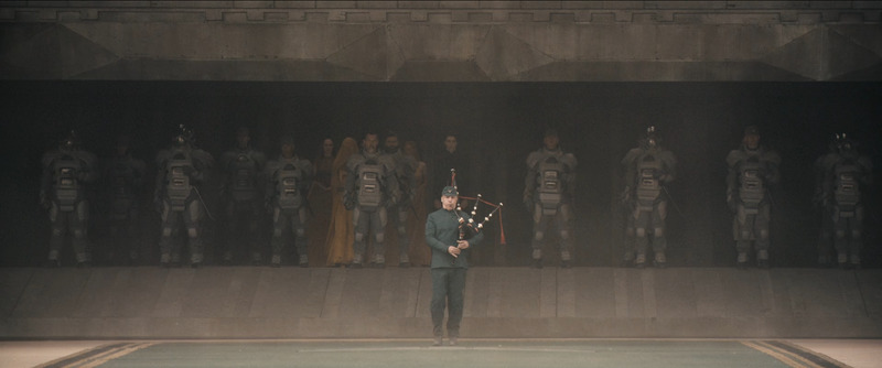 An Atreides musician plays the bagpipes, backed by a battalion of Atreides warriors obscured by the shadow of the space ship in the background.