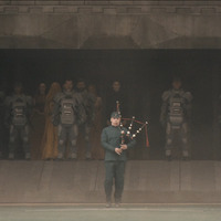 An Atreides musician plays the bagpipes, backed by a battalion of Atreides warriors obscured by the shadow of the space ship in the background.
